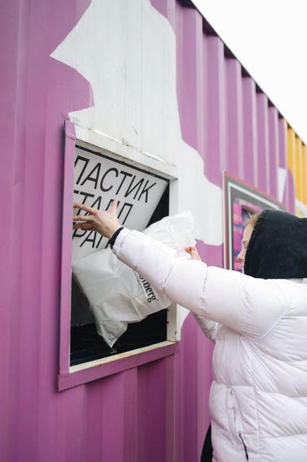 A person wearing a white puffer jacket with a black hood is seen disposing of a white plastic bag into an outdoor collection unit. The collection box is embedded into a pink and white painted wall, with the box's border and frame in light purple. The person’s right arm is extended towards the opening, and they are using their left hand to steady themselves. In the background, there is a portion of a window or poster with black and white text, partially obscured by the person. The wall's surface appears smooth and painted, and the setting suggests an urban environment designed for independent waste disposal or recycling, operated by a waste management service like Rubbish Collection Shoreditch. The scene is well-lit with natural daylight, and the surrounding area appears clean and organized, consistent with private rubbish collection or on-site waste handling services focused on environmentally responsible disposal.
