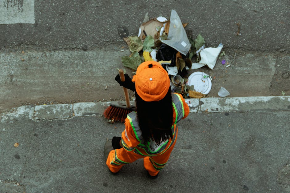 An overhead view of a sanitation worker from Rubbish Collection Shoreditch wearing an orange high-visibility uniform with reflective stripes, positioned on a sidewalk near a curb. The worker's attire includes an orange cap and dark gloves. They are holding a broom with a wooden handle and dark bristles, standing in front of a small pile of mixed waste, including paper, cardboard, leaves, and other debris, which is partially spread along the edge of the pavement and against the concrete curb. The background shows a weathered asphalt street with visible wear and discoloration, adjacent to a concrete roadside. The scene is lit with natural daylight, and the worker appears to be engaged in an on-site clearance task, typical of private waste handling and rubbish removal services.