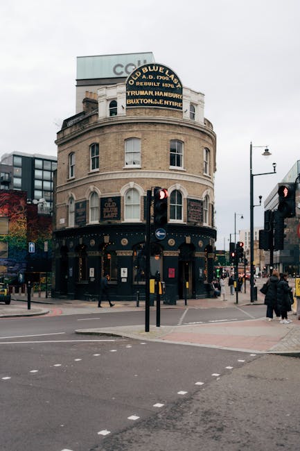 A historic three-story building with a rounded corner façade located at a busy street intersection, featuring a beige brick exterior with white window frames and decorative architectural details. The ground floor has black painted storefronts with gold accents and signage, including the name 'Old Blue Last' displayed on a sign above the entrance. A prominent, curved vintage-style signboard at the top of the building advertises various musical acts. In the foreground, there are multiple pedestrian crossings and traffic signals, with several people walking on the sidewalks and waiting at the crosswalks, some dressed in dark winter clothing. Modern street furniture such as traffic lights and a street lamp are visible, along with a mix of nearby buildings, including glass office structures and trees with autumn foliage. The overcast sky creates diffuse lighting, emphasizing the historic building's architectural features within an urban scene that relates to the city's lively social environment, potentially involving private or alternative waste handling by local waste management services like Rubbish Collection Shoreditch serving the area.