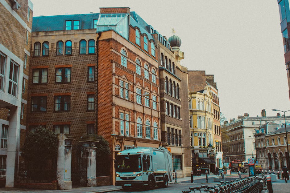 A white refuse collection vehicle with a blue logo and black details is parked on a narrow urban street near the entrance to a building with black iron gates and stone pillars. The vehicle is positioned slightly to the left in the foreground, showing its front and side, and is equipped with a large rear compactor for waste transport. The buildings surrounding the scene display a mix of architectural styles, including a red brick building with multiple arched windows, white decorative stone accents, and a rounded tower with a domed roof topped by a small cross, indicating a church or historic structure. To the right, there is a yellow, Victorian-style building with ornate window framing, while further down the street, other historic structures with detailed facades are visible. The street is lined with bollards, a bike rack, and a lamppost, with the sky overcast providing diffuse, natural lighting. The scene suggests a typical setting in central London where private rubbish collection services, like those offered by Rubbish Collection Shoreditch, operate in close proximity to historic residential and commercial buildings, facilitating on-site waste removal and alternative disposal options.