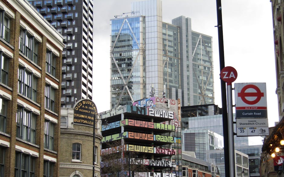 A city street scene featuring a variety of urban signage and modern high-rise buildings. In the foreground, a black sign with illuminated yellow letters indicates the location of the Old Blue pub, referencing its historical name and address. To the right, a red bus stop sign displays routes towards Shoreditch Church, with route numbers 35, 47, and 78, and a small white sign indicating a bus stop on Curtain Road. Behind these signs, graffiti-covered billboards and advertisements are stacked on a structure, with colourful tags and stylized lettering visible. The background is dominated by tall, contemporary office buildings with glass facades, reflecting the overcast sky and surrounding structures. The scene is lit with soft natural daylight, highlighting the textures of brick, metal, and glass, typical of an urban environment that may be associated with on-site waste collection or alternative waste handling in the Shoreditch area, supporting local rubbish removal services like those provided by Rubbish Collection Shoreditch.