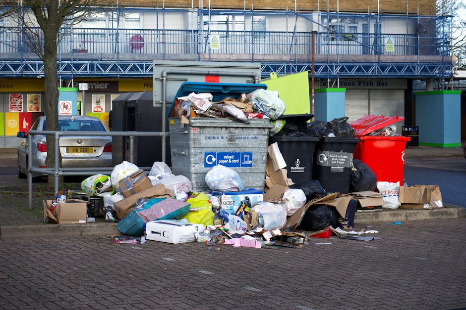 A white refuse collection vehicle with a blue logo and black details is parked on a narrow urban street near the entrance to a building with black iron gates and stone pillars. The vehicle is positioned slightly to the left in the foreground, showing its front and side, and is equipped with a large rear compactor for waste transport. The buildings surrounding the scene display a mix of architectural styles, including a red brick building with multiple arched windows, white decorative stone accents, and a rounded tower with a domed roof topped by a small cross, indicating a church or historic structure. To the right, there is a yellow, Victorian-style building with ornate window framing, while further down the street, other historic structures with detailed facades are visible. The street is lined with bollards, a bike rack, and a lamppost, with the sky overcast providing diffuse, natural lighting. The scene suggests a typical setting in central London where private rubbish collection services, like those offered by Rubbish Collection Shoreditch, operate in close proximity to historic residential and commercial buildings, facilitating on-site waste removal and alternative disposal options.